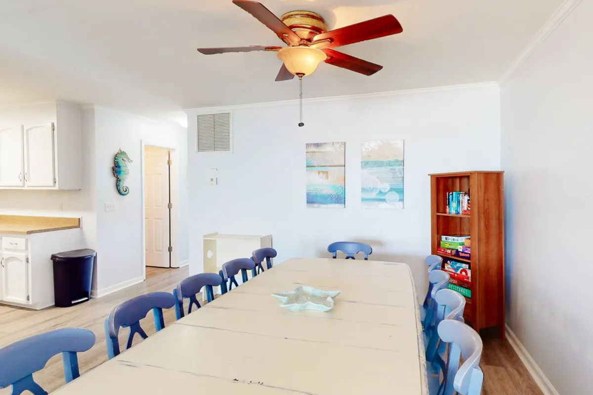A spacious dining area featuring a long table surrounded by blue chairs, a ceiling fan overhead for comfort, wall art, and a bookshelf hinting at leisure and family moments. The arrangement suggests readiness for group meals, celebrations, or game nights.