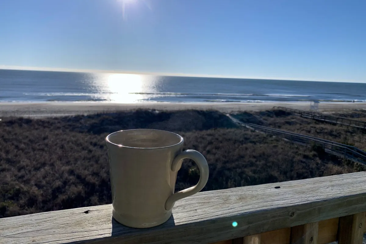 A relaxing deck scene with a coffee mug in the foreground, sunlight sparkling on the ocean, and gentle waves in the distance. The mood is serene, capturing the peaceful moments of sunrise or quiet reflection by the sea.