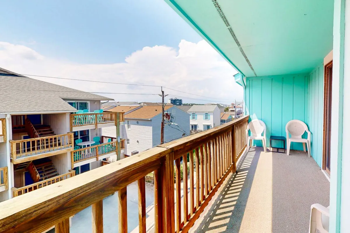 A casual street-side balcony with two plastic chairs and a wooden railing, overlooking nearby buildings. The setup is ideal for enjoying morning coffee, people-watching, or soaking in the breeze. The street view adds a lively, connected-to-town atmosphere, contrasting with the seclusion of the oceanfront deck.