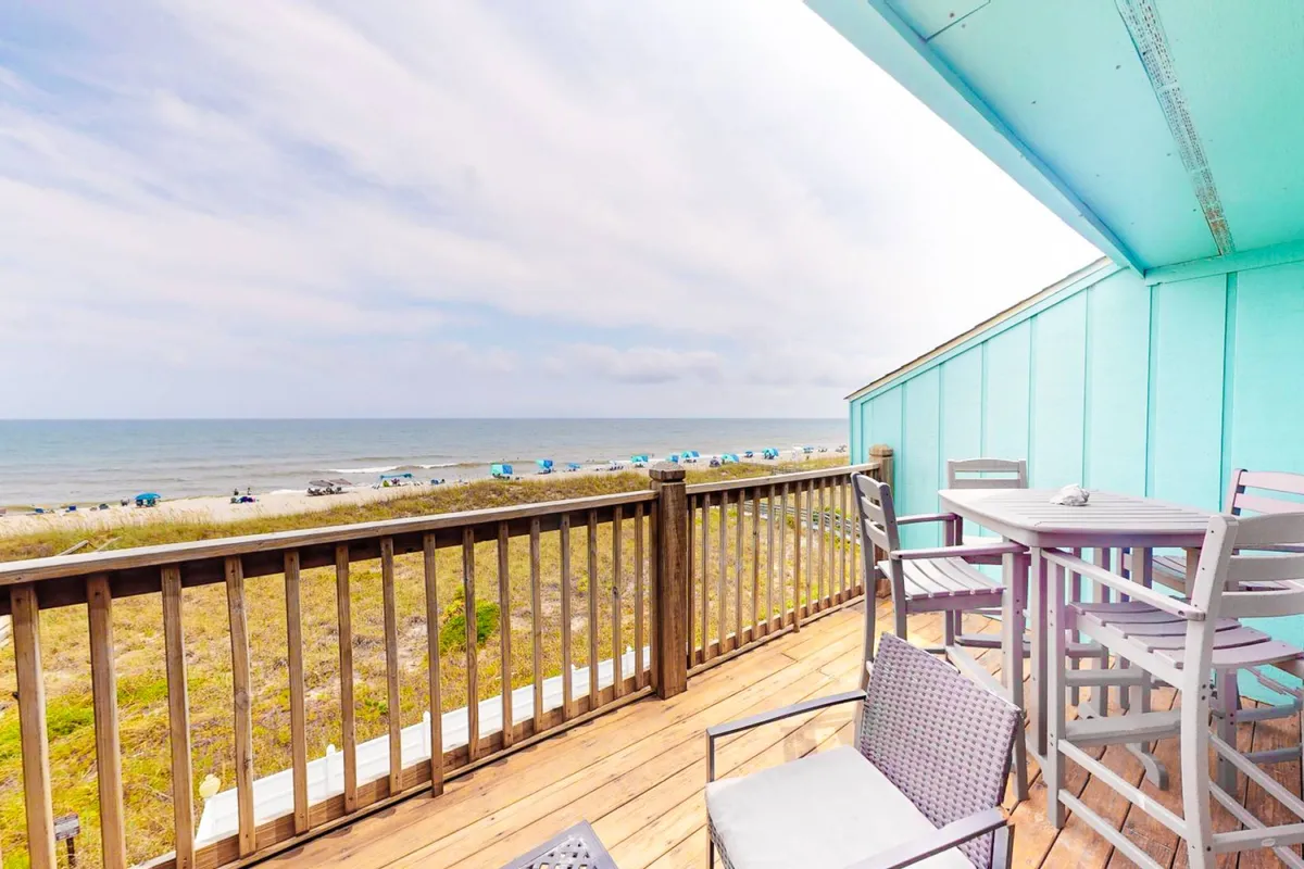 This image shows a serene oceanfront deck with comfortable outdoor furniture and sweeping views of the water and beach. The deck is bathed in soft morning or evening light, creating an inviting space for relaxation or social gatherings. Deck furniture is arranged to encourage both lounging and conversation, while the beach umbrellas in the distance reinforce the coastal setting.