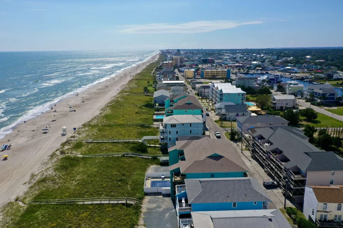 A vibrant exterior shot capturing the sandy beach, sparkling ocean, residential buildings, and a lively boardwalk area populated with people. The image conveys a sense of proximity to the beach and the social attractions of the Carolina Beach area.