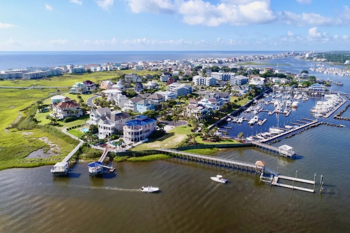 A sweeping view of luxurious waterfront homes, a bustling marina with boats, expansive green spaces, and a lively coastal landscape. The mood is relaxed and vibrant, with evidence of boating activities and community life along the shore.