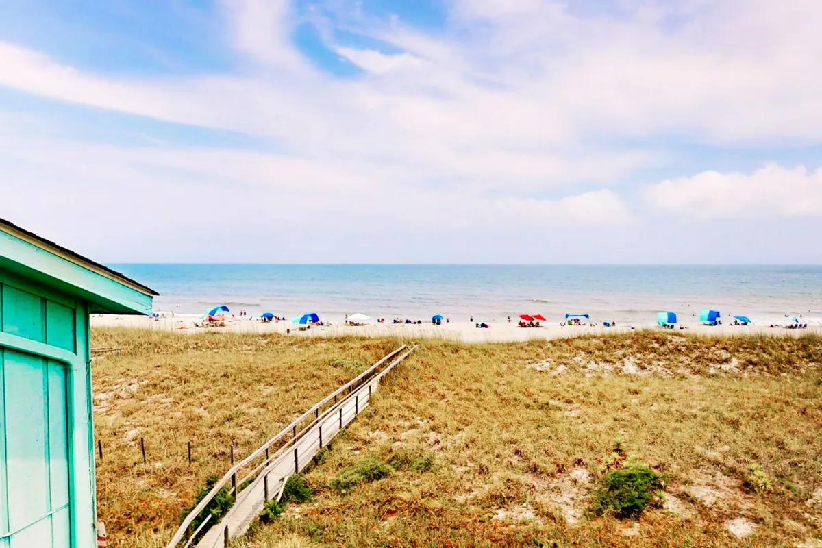 A stunning oceanfront view with golden sand, turquoise water, beach umbrellas, and the boardwalk visible in the distance. The scene is lively yet tranquil, inviting visitors to enjoy everything from beach games to sunset strolls just steps from the property.