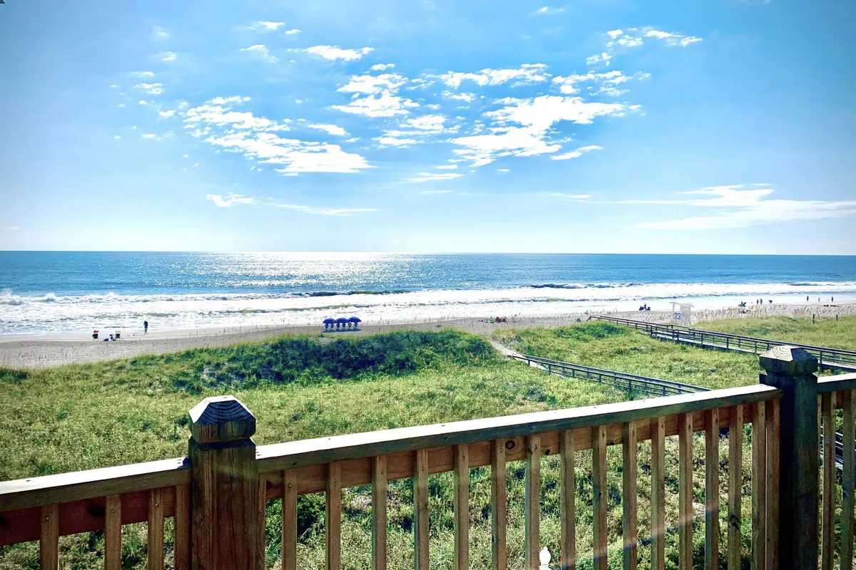 A panoramic view from the property’s deck, showing the pristine sandy beach, gentle ocean waves, lush dune vegetation, and a sturdy wooden railing. The tranquil sky and open space evoke endless possibilities for family adventures and relaxation.