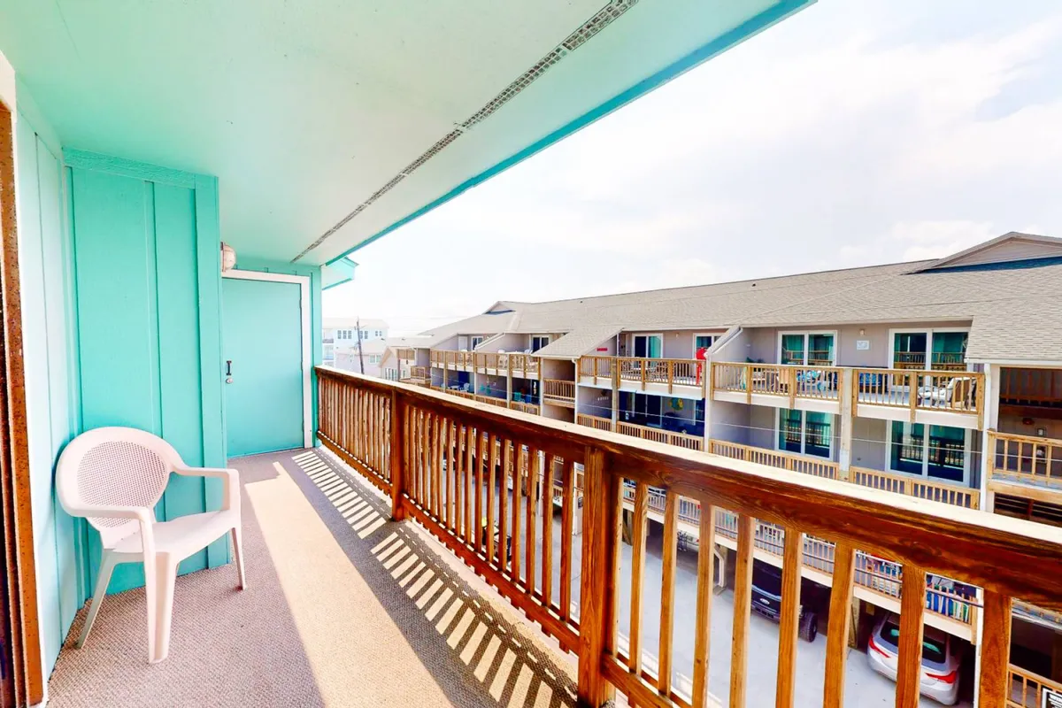 A relaxed exterior view showing a condo deck with a sturdy wooden railing, a plastic chair positioned for comfort, and a turquoise wall. The scene suggests a casual spot to unwind, chat, or sip drinks while enjoying the beach breeze.