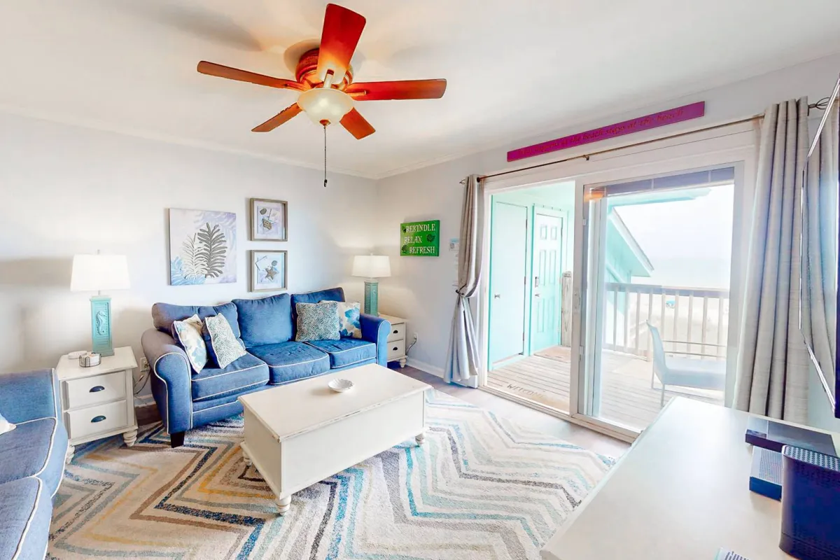 This interior scene captures a blue sofa adorned with colorful throw pillows, a coffee table ready for board games or drinks, a decorative wall art accent, ceiling fan, and a sliding glass door that opens directly onto the deck. Sunlight streams in, creating a bright, open-space feel while emphasizing the seamless indoor-outdoor connection.