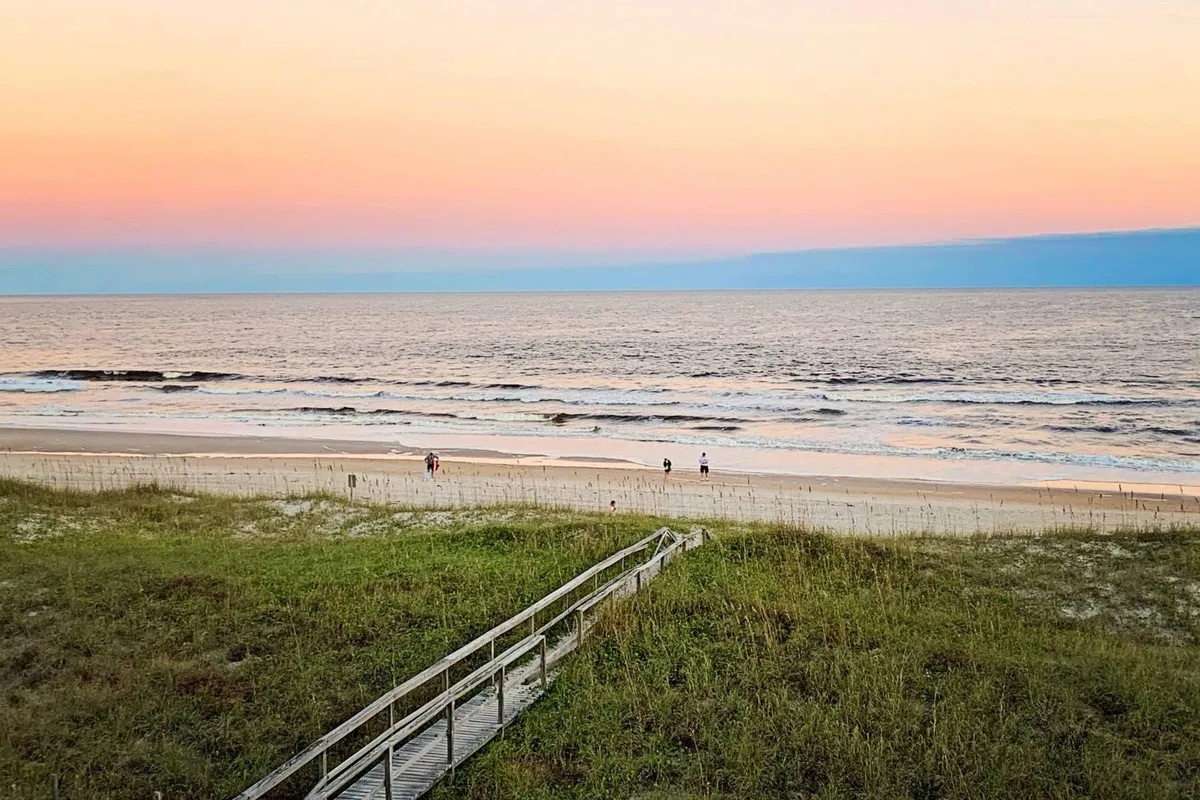 The image showcases a stunning, unobstructed view of the ocean at sunset, with the golden sky reflected on the water. A wooden boardwalk leads directly to the sandy beach, where people are walking and families are gathered, evoking a sense of togetherness and relaxation. The scene captures both the serene beauty of the Carolina Beach coastline and the inviting beach access that the Sea Turtle's Nest condo offers.