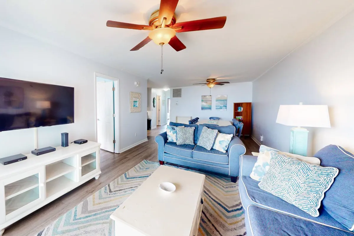 A cozy living room setup with a plush sofa adorned with decorative pillows, a coffee table ready for games or snacks, and a TV unit, all under a ceiling fan. The atmosphere is casual and perfect for shared activities like movie nights or family games.