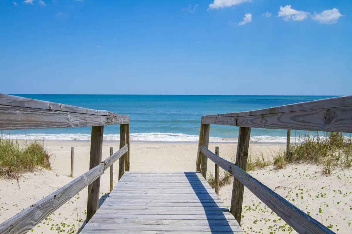A sunlit wooden boardwalk stretches from the condo complex through sandy dunes, leading directly to the beach and ocean under a wide blue sky. The inviting path signals private and easy beach access for guests.