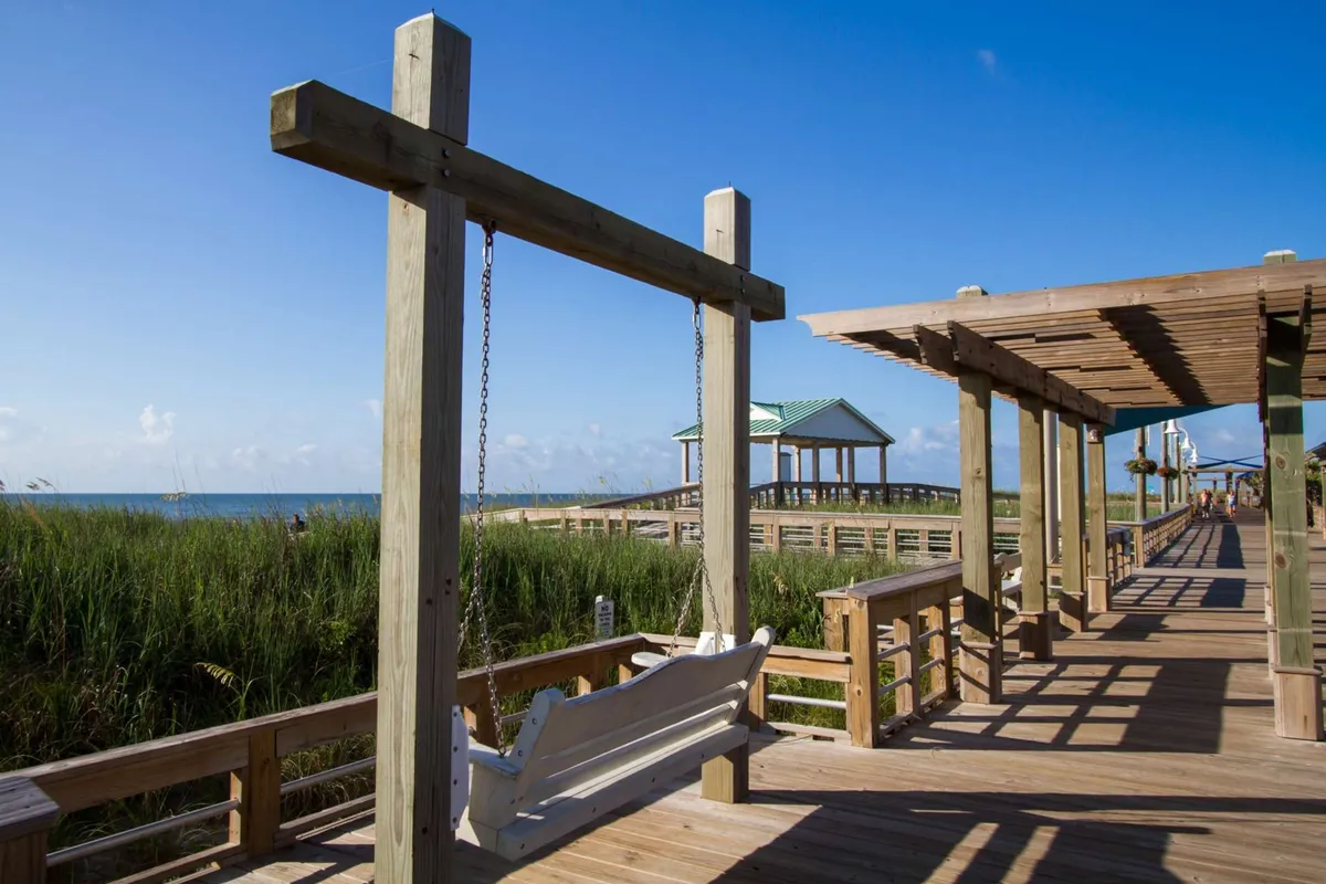 A serene exterior view of a wooden swing on a grassy area overlooking the boardwalk and the ocean, with pavilions in the background. The inviting swing and panoramic setting suggest a peaceful spot for gathering, conversation, and sunset watching.