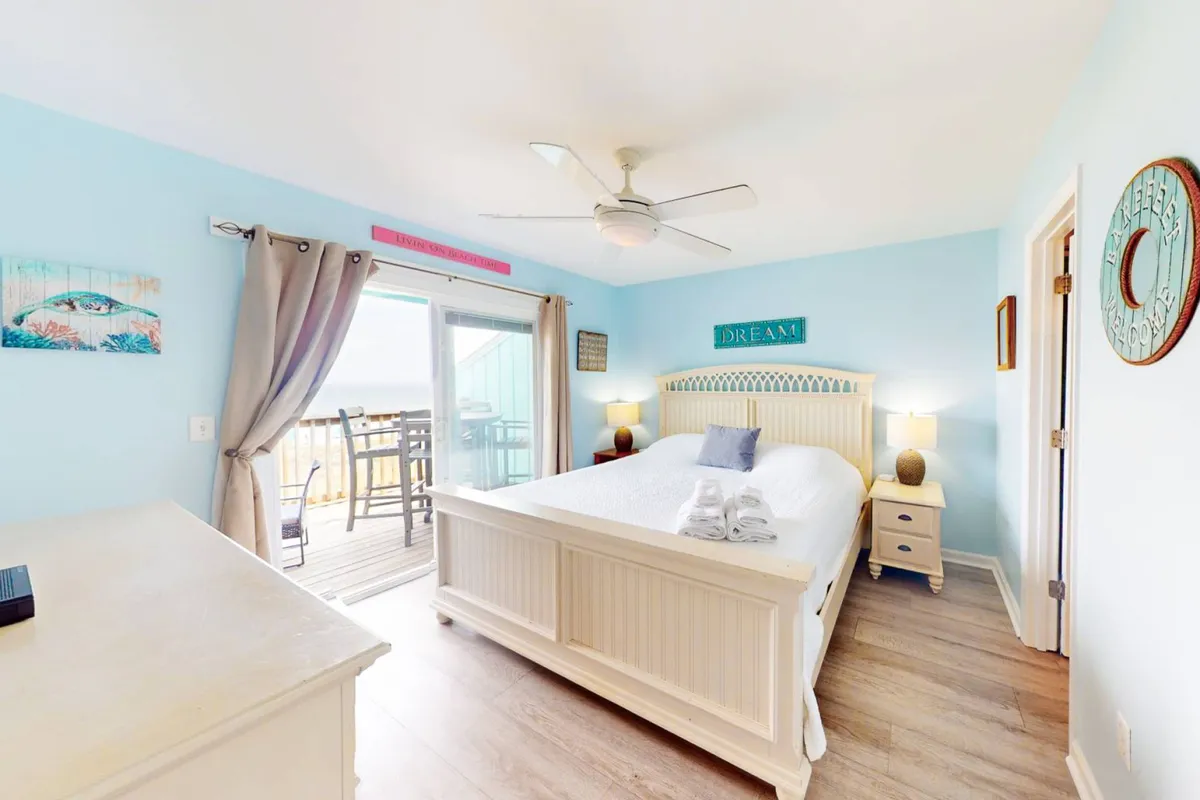 A tranquil bedroom scene with a plush bed facing a balcony door that opens to expansive ocean views. Decorative signs and soft natural light set a relaxing, upscale tone, illustrating the blend of private comfort and outdoor connection.