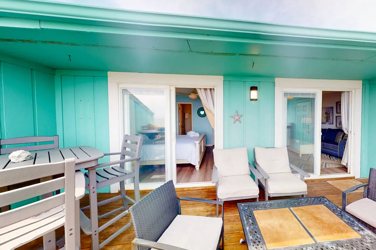 A welcoming outdoor seating area framed by turquoise walls, with a coffee table and sliding glass doors that offer a glimpse into the interior. The area is arranged for relaxation or socializing, perfect for al fresco meals or sunset gatherings. The mood is casual and inviting, blending indoor and outdoor living.